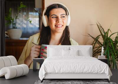 One young girl or woman is listening to music on her wireless headphones while drinking her morning coffee or tea in the house before going to work Wall mural