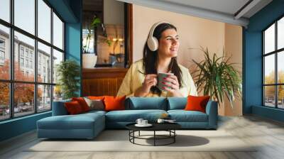 One young girl or woman is listening to music on her wireless headphones while drinking her morning coffee or tea in the house before going to work Wall mural