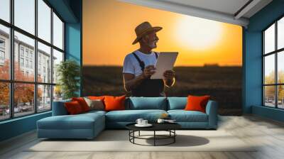 An elderly man stands in a field being plowed, holding a digital tablet that helps him monitor farm machinery. In the background is the sunset. Wall mural