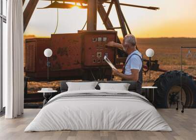 Older farmer holding tablet in his hands and adjusts irrigation system on field at sunset. Wall mural