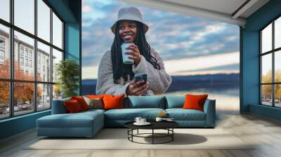 Portrait of a beautiful black woman smiling excitedly at the camera, she's wearing a bucket hat and a backpack, she's drinking coffee and taking pictures with her phone Wall mural