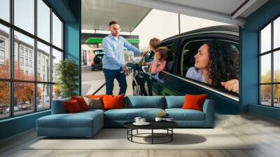 The father fills the fuel and talks to his daughters who are sitting in the back seat of the car, he is upset because they did unfasten their seat belts. Mom and daughters look at father Wall mural