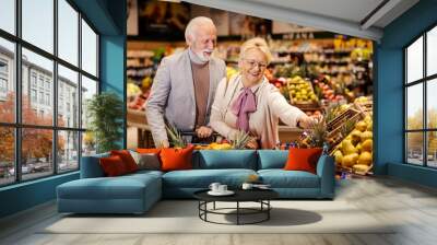 A cheerful senior couple is purchasing fresh, healthy groceries at the supermarket. Couple is buying fruits. Wall mural