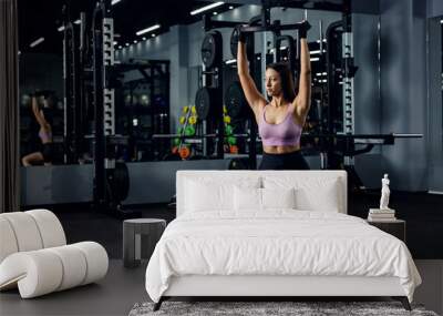 A fit woman in sportswear is doing squats in the gym with a dark atmosphere. She wears training gloves and holds a heavyweight over her head. Body shape, stay in Wall mural