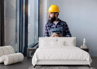 A happy, bearded worker with a helmet on his head, is standing next to a window and using his phone to check when is material arriving. He is looking through the window. Wall mural