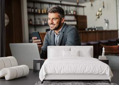 A happy businessman is sitting in a coffee shop and checking on his bank account on the mobile. There is a laptop on a table. A man using the phone for e-banking Wall mural