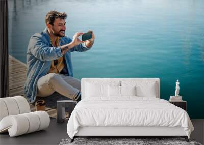 A happy man is sitting on a dock and taking selfies. Wall mural