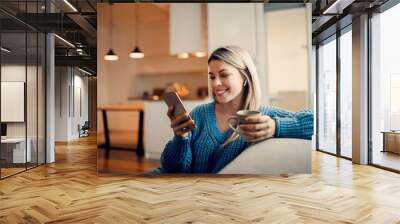 A happy woman hanging on social media on the phone and drinking coffee at home. Wall mural