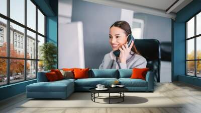 A smiling businesswoman talking on the phone with client, using computer and multitasking at office. Wall mural