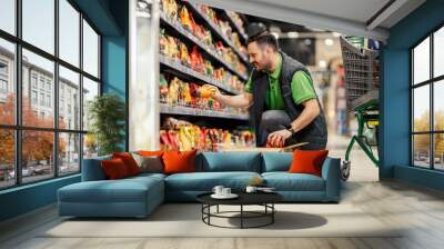 A stock clerk is crouching and displaying groceries on shelves at supermarket. Wall mural