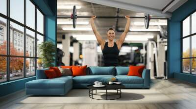 A strong happy woman achieved goal. A female bodybuilder lifted barbell in the air in a gym. Wall mural