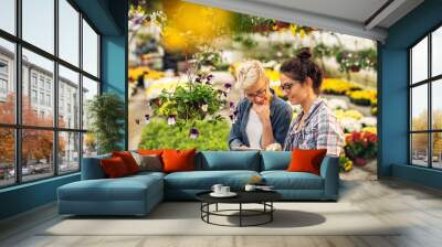 Adorable charming professional modern florist woman showing a list of flowers on a tablet to the curious attractive blonde female customer in a greenhouse. Wall mural