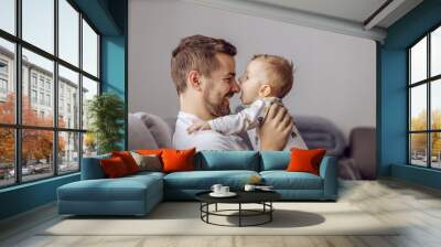 Adorable little blond boy playing with his caring father and trying to bite his nose. Family values. Wall mural