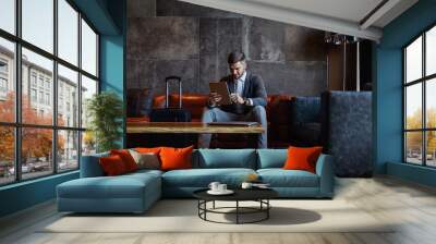 An elegant handsome man is sitting in the hotel lobby and using a tablet. He is on a business trip and carries around his hand luggage and laptop. Travel, business trip, away from home, online Wall mural