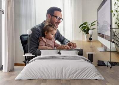 An entrepreneur is typing a report while holding his daughter in his lap at home office. Wall mural