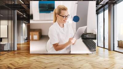 Attractive blond lab assistant in sterile uniform with eyeglasses sitting and using tablet for entering test results. Wall mural