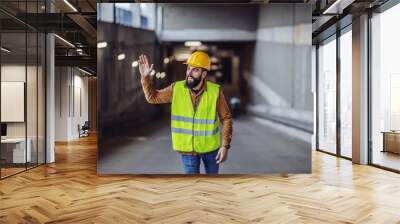 Attractive cheerful bearded friendly worker in vest, with helmet on head exiting building in construction process, waving and saying hello to other workers. Wall mural