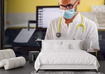 Attractive male scientist with face mask and rubber gloves standing in laboratory and holding test results in hands. Corona virus outbreak concept. Wall mural