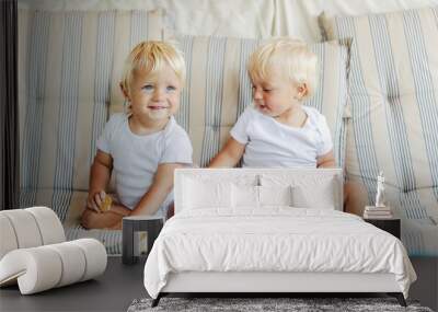 Babies eating cookies and playing around. 2 happy toddlers in white baby bodies holding a cake in their hand on a beige striped sofa One child smiles and looks away while the other looks at a relative Wall mural