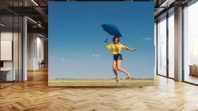 Beautiful Caucasian girl in yellow blouse and denim shorts jumping with blue umbrella outdoors. Blue sky with clouds in background. Wall mural