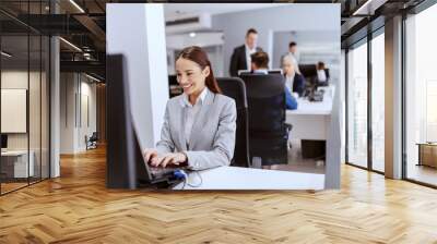 Beautiful positive Caucasian businesswoman in formal wear sitting in office and using computer. Hands on keyboard. In background colleagues working. Wall mural