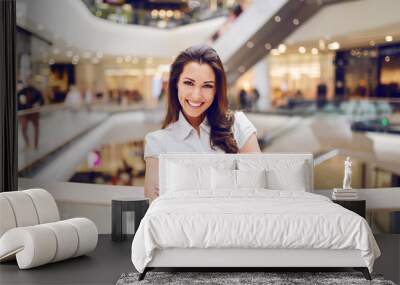 Beautiful smiling caucasian brunette in shirt standing in shopping mall with arms crossed. Wall mural