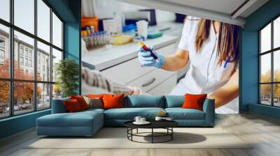 Caucasian laboratory assistant in uniform, with mask and rubber gloves holding test tube with blood while patient sitting and holding absorbent cotton. Wall mural