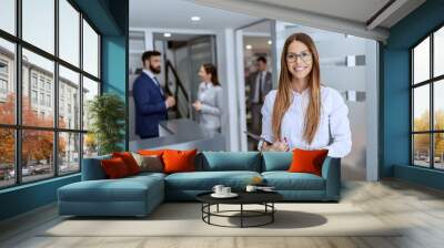 Charming Caucasian businesswoman dressed in shirt and skirt standing on hallway and holding tablet. In background her colleagues standing and chatting. Wall mural