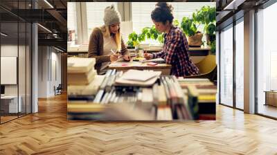 Charming stylish beautiful hipster student female friends sitting in the sunny school library near the window and doing homework together with notes and a tablet. Wall mural