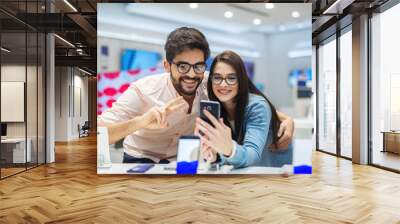 Cheerful beautiful young couple in bright big electronic store testing new telephone. Shopping together and having fun. Wall mural