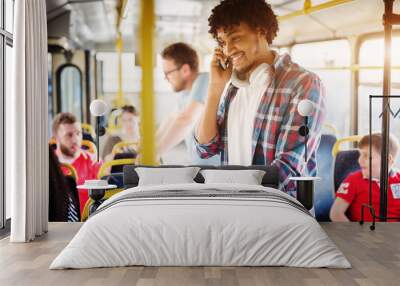 Cheerful young man with headset hanging around his neck is talking on a phone and smiling while standing on a bus. Wall mural