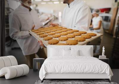 Close up of fresh cookies on big tray in food factory. Blurred picture of two male employees in sterile clothes talking in background.One man is holding tray. Wall mural