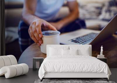 Close up of man taking mug with morning coffee from desk. Man is dressed in pajamas, sitting on sofa in living room and using laptop. Weekend activities. Selective focus on hand. Wall mural