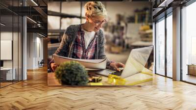 Close up view of charming smiling motivated short hair attractive middle aged industrial female engineer with eyeglasses working with blueprints and laptop in the workshop. Wall mural