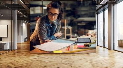 Confident serious attractive professional architect woman leaning against the desk and looking in the new project with a notes, tablet, and rulers on the table in the fabric place. Wall mural