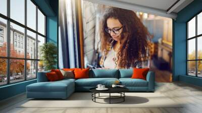 Cute mixed race girl with curly hair standing in cafeteria and using smart phone. Wall mural