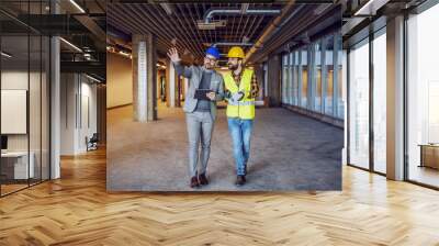 Dedicated innovative architect holding tablet and explaining his ideas to construction worker. Building in construction process interior. Wall mural