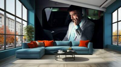 Elegant modern businessman with laptop on the back seat talking on a mobile. Wall mural