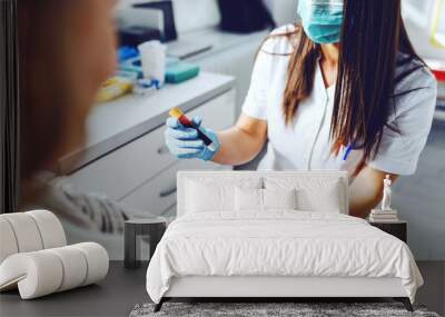 Female lab assistant in uniform, with protective mask and rubber gloves holding test tube with blood and holding patient's arm. Patient holding cotton on vein. Wall mural