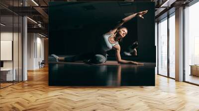 Fit young Caucasian brunette in sportswear sitting on mat in gym and doing side stretching. In background mirror. Wall mural