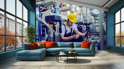 Focused plant worker in overalls, with protective helmet on head and antiphons on ears using tablet for checking machine while crouching. Wall mural