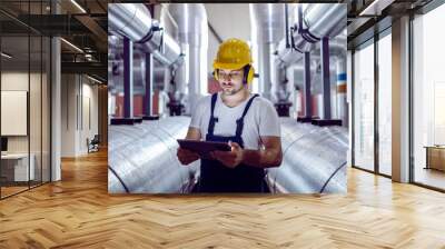 Focused plant worker in overalls, with protective helmet on head and antiphons on ears using tablet for checking machine. Wall mural