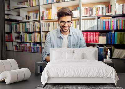 Front view of young hipster smiling man reading a book on the library floor. Wall mural