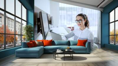 Gorgeous smiling businesswoman with brown curly hair, ponytail and eyeglasses dressed casual sitting at office and using laptop. Wall mural