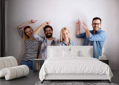 Group of young handsome colleague people standing against the wall showing at empty editable space above while one girl standing with thumb up and looking at the camera. Wall mural