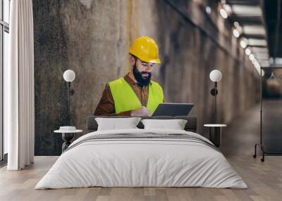 Handsome bearded supervisor in vest with helmet on head standing in building in construction process and using laptop to check is everything according to plan. Wall mural