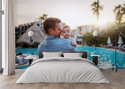 Handsome Caucasian dad hugging son and looking at him while standing next to swimming pool. Baby looking over father's shoulder. Wall mural