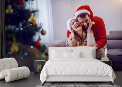 Handsome caucasian man giving christmas present to his loving girlfriend. Woman hugging man and thanking him for gift. Both are dressed in sweaters and having santa hat on heads. Christmas holidays. Wall mural