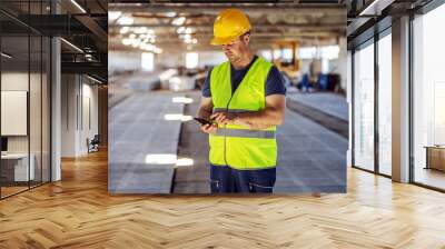 Hardworking blue collar worker standing at construction site and using smart phone to order some more materials for building. Wall mural