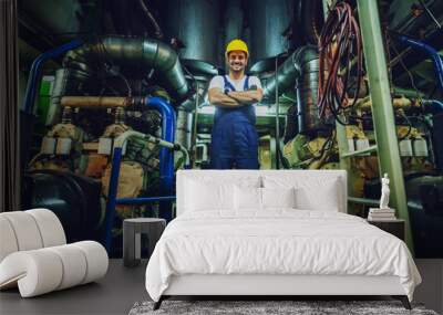 Low angle view of handsome caucasian worker in overall and helmet on head standing in ship next to engine with arms crossed. Wall mural